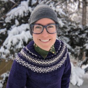 a smiling photo of Maia in the wintertime. She has light skin and blue eyes, and she is wearing an indigo and white hand-knitted sweater, a collared shirt, a grey knit cap, and large blue-framed glasses. She is smiling and leaning toward the camera, and there is snow covering the trees in the background.